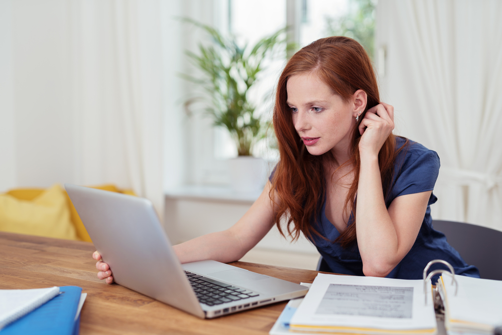 Pretty young student studying at home sitting at her dining table with a large binder of notes checking something on the screen of her laptop computer